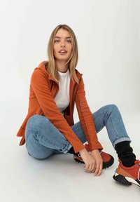 Rostorange, enganliegende Zip-Jacke mit Kapuze, kombiniert mit einem weißen T-Shirt und hellblauen Jeans. Rote und schwarze Sportschuhe mit weißen Schnürsenkeln.