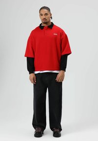 Rood oversized poloshirt met een zwarte kraag, gedragen over een zwart shirt met lange mouwen. Gecombineerd met zwarte jeans met wijde pijpen en zwarte schoenen.