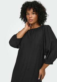 Melna kleita ar brīvu piegriezumu, apaļu kakla izgriezumu un apjomīgām puff piedurknēm. Izgatavota no teksturēta auduma ar smalkām šuvju detaļām.