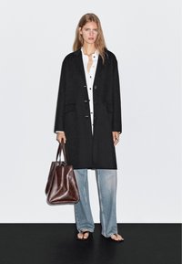 Czarny, oversize płaszcz z guzikami, noszony na białej koszuli, w połączeniu z jasnoniebieskimi spodniami typu wide-leg oraz brązową skórzaną torbą.