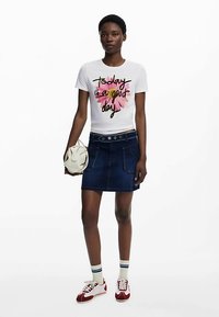 T-shirt blanc avec un motif floral coloré et un texte noir, associé à une jupe en jean. Accessoires : un sac crème et des baskets rouges.