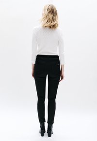 Zwarte skinny jeans met achterzakken en een gladde textuur, gecombineerd met een witte gebreide trui. Model gezien van achteren, draagt zwarte laarzen.
