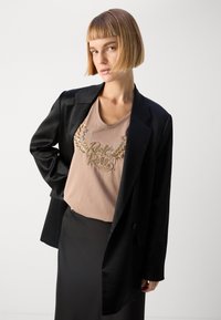 Černý blazer přes béžové tričko se zlato-stříbrnými ozdobami znázorňujícími křídla. Viditelná černá sukně, celkově hladká textura.