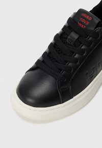 Baskets en cuir noir avec semelle blanche épaisse, lacets noirs et texte rouge "HUGO YOUR WAY" sur la languette.