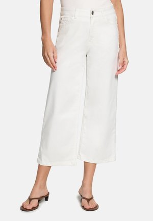 Kvinde iført hvide, vidde, cropped bukser og brune sandaler med hæl, stående foran en ensartet lys baggrund.