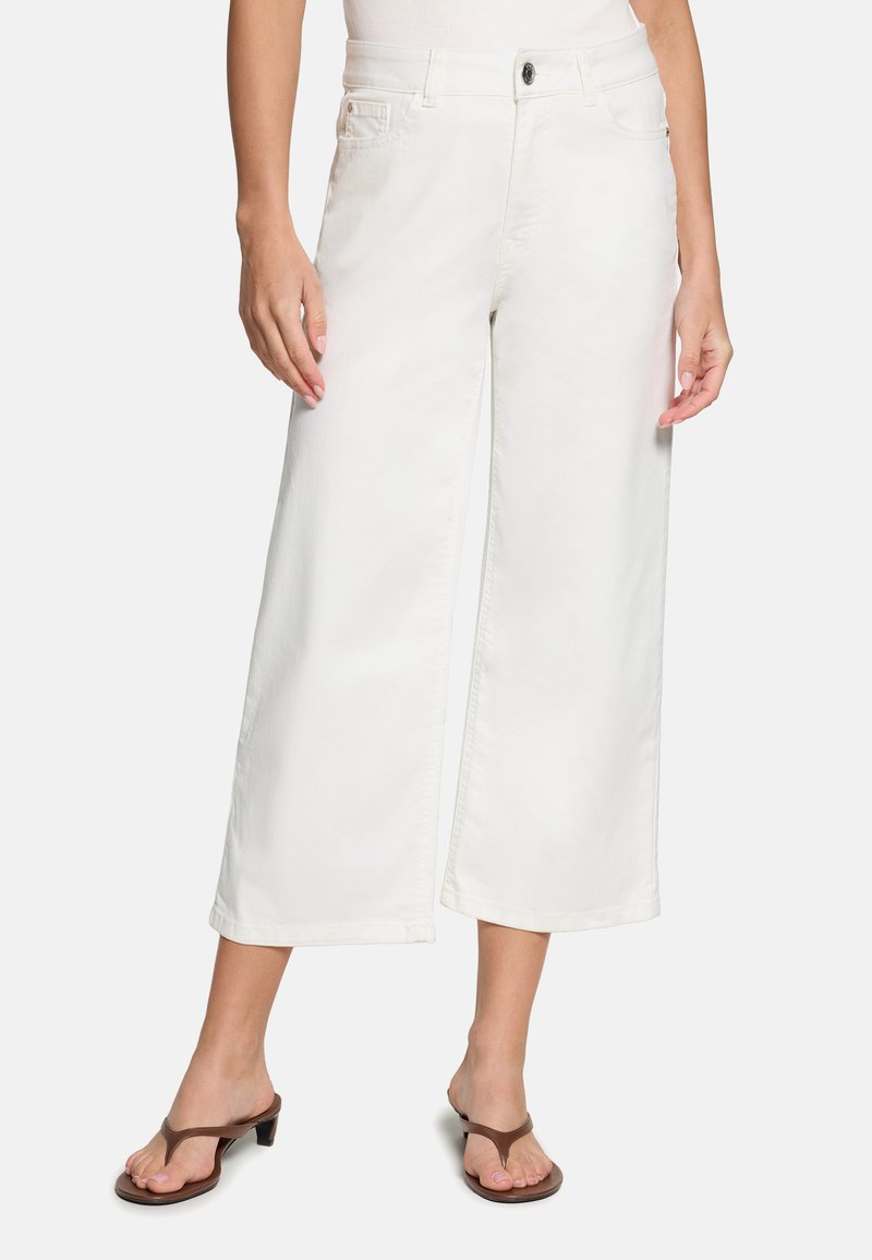 Femme portant un pantalon blanc large et court et des sandales marron à talons, debout devant un fond clair uni.