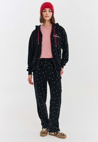 Čierna mikina s bodkami a červenými šnúrkami, spárovaná s čiernymi nohavicami. Svetloružová košeľa dole dotvára ležérny outfit.