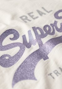Cremefarvet stof med sølv- og glitrende lilla tekst, der læser "REAL Super", sammen med dekorative scroll-detaljer.