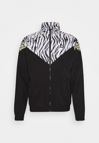 Chaqueta negra con cremallera, parte superior con estampado de cebra blanco, acento de leopardo dorado en el hombro y puños elásticos. Tejido ligero, diseño deportivo.