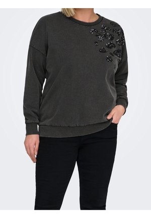 Kobieta ubrana w ciemnoszarą bluzę z długim rękawem z czarnymi cekinowymi kwiatowymi zdobieniami na lewym ramieniu oraz czarne spodnie, stojąca z ręką w kieszeni.
