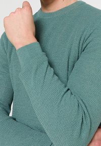 Suéter color verde agua claro con un patrón de punto texturizado, cuello redondo y puños acanalados. La tela parece suave y ligeramente elástica.