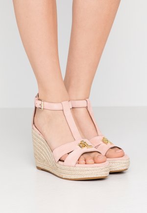 Feet wearing pink suede wedge sandals with ankle straps and gold logo detail on front over woven platform soles.