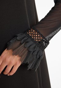 Détail d'une robe noire avec une manche noire transparente aux bords festonnés et un accent en maille au niveau du poignet, mettant en avant la texture et les éléments de design.