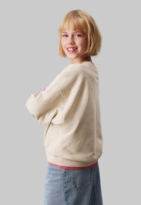 Sudadera de color crema con un ajuste relajado, puños y dobladillo acanalados. Combinada con jeans de mezclilla azul claro. Textura plana y diseño casual.