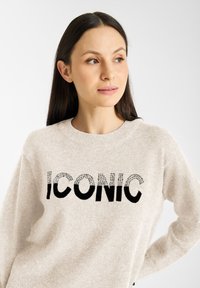 Jasno beżowy sweter z wyraźnym czarnym napisem "IKONICZNY", ozdobiony małymi srebrnymi detalami. Miękka tekstura i okrągły dekolt.