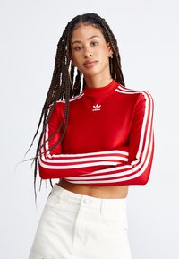 Κόκκινο μακρυμάνικο μπλουζάκι από ελαστικό υλικό, με λευκές τρεις λωρίδες κατά μήκος των μανικιών και ένα μικρό λογότυπο στο στήθος.