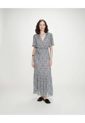 Femme debout portant une robe maxi fleurie bleu-gris ceinturée, à manches courtes, avec des chaussures plates noires sur un fond blanc uni.