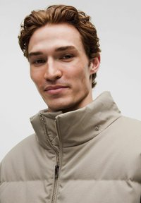 Junger Mann mit heller Haut und welligem braunem Haar, der eine beige gesteppte Jacke mit hohem Kragen trägt und mit einem leichten Lächeln in die Kamera schaut.