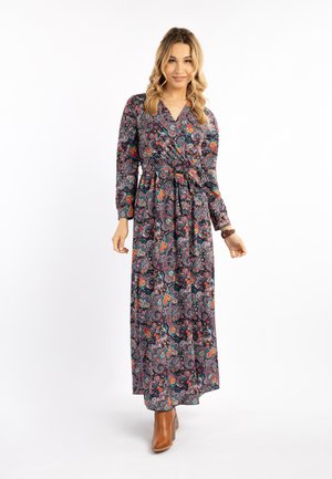 Femme portant une robe à manches longues, longue jusqu'aux chevilles avec un motif floral, et des bottines marron, debout sur un fond blanc.