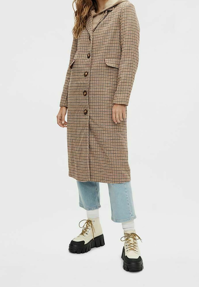 Brun lång rock i hundtandsmönster med stora knappar, två sidfickor och krage, kombinerad med ljusblå korta jeans och kraftiga stövlar.