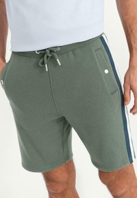 Pantaloni corti in cotone verde con vita regolabile, tasche laterali e strisce blu navy sui lati. Texture morbida con vestibilità elastica.