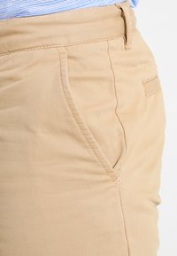 Pantalon khaki en coton lisse, doté d'une taille plate, de poches latérales et d'une coupe slim. Les coutures sont visibles le long des bords.