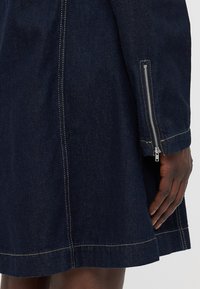 Temno jeans obleka z razširjenim dizajnom, vidnim kontrastnim šivanjem in izpostavljenim zadrgo na levem rokavu.