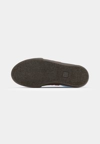 Semelle d'une chaussure marron avec une adhérence texturée et un petit logo rectangulaire près du talon sur un fond clair uni.