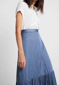 Kvinna iförd en vit kortärmad knapp-up blus och en högmidjad, plisserad blå midi kjol mot en enkel bakgrund.