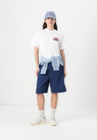 Balta trumpa rankovė t-shirt su raudonu grafiku, mėlynas džemperis surištas ant liemens, tamsiai mėlyni šortai ir šviesiai pilki batai su smėlio spalvos akcentais.