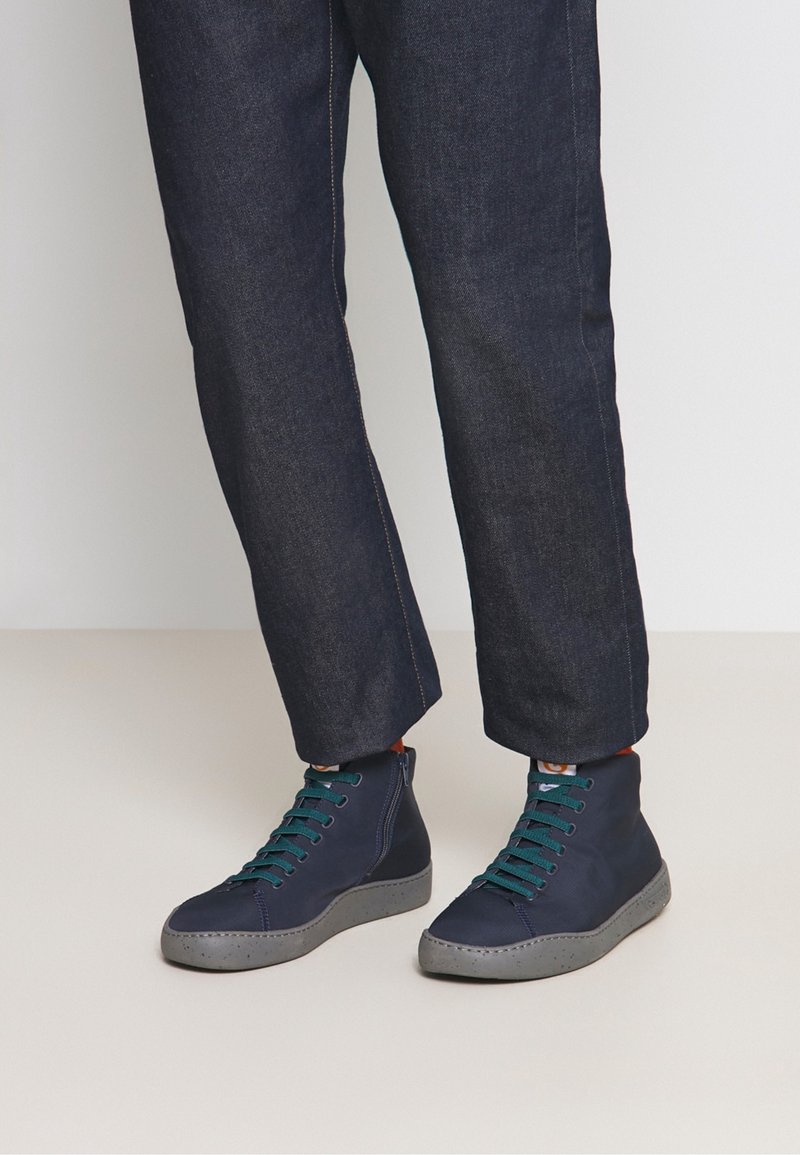 Personne portant des baskets montantes bleu foncé avec des lacets verts et un pantalon en jean foncé, debout sur un sol clair.