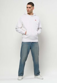 Hombre con sudadera blanca con capucha y logo rojo, vaqueros azules y zapatillas blancas, de pie con las manos en el bolsillo de la sudadera, mirando hacia la izquierda.