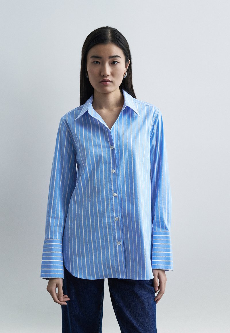 Γυναίκα με μακριά σκουρόχρωμα μαλλιά φοράει μπλε και λευκό πουκάμισο με κάθετες ρίγες και φαρδιές μανσέτες, καθώς και σκούρο παντελόνι, στέκεται μπροστά σε απλό φόντο.