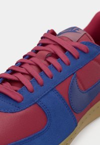 Røde og blå sneakers med en tekstureret stofoverdel, der har matchende snørebånd, et blåt Nike-logo og en beige gummisål.