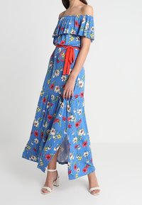 Frau in einem blauen, schulterfreien, floralen Maxikleid mit einem roten Gürtel und weißen High-Heels, die seitlich vor einem weißen Hintergrund steht.