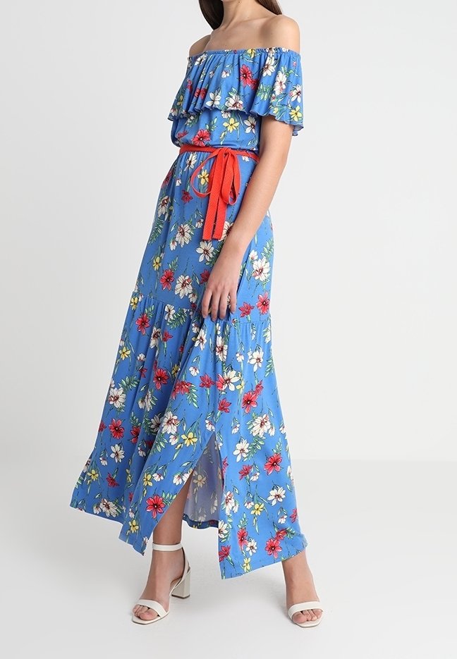 Frau in einem blauen, schulterfreien, floralen Maxikleid mit einem roten Gürtel und weißen High-Heels, die seitlich vor einem weißen Hintergrund steht.