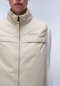 Gilet matelassé beige avec un col montant, doté d'une fermeture éclair et de coutures décoratives. Design minimaliste avec un logo brodé sur la poitrine.