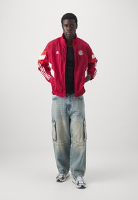 Giacca rossa con zip, dettagli bianchi e logo, maglietta nera, pantaloni cargo azzurri larghi e sneakers bianco e nero.