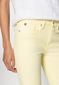 Jeans de mezclilla amarillos con un corte slim. Cuenta con un bolsillo delantero, herrajes plateados y un pequeño acento tricolor en el bolsillo. La textura es suave.