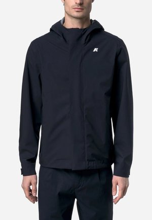 Uomo che indossa una giacca nera aderente con cappuccio e un piccolo logo bianco sul petto, in piedi davanti a uno sfondo uniforme.