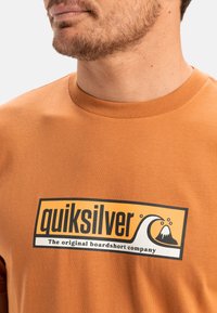 Camiseta de algodón naranja con un gran logo en blanco y negro de "Quiksilver" que presenta un gráfico de olas y montañas en la parte frontal. Diseño de cuello redondo.