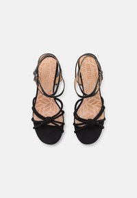 Le sandali in suede nero presentano molteplici cinture incrociate e un design annodato. Le suole interne sono in pelle di colore chiaro con un motivo a foglia.