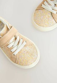 Baskets beige clair avec une surface texturée ornée de paillettes iridescentes. Lacets et bande blanche. Semelles en caoutchouc avec un bord arrondi.