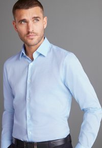 Himmelblaues, langärmliges Hemd mit Knopfleiste, subtiler Textur, spitzem Kragen und weißen Knöpfen; getragen mit einem dunklen Gürtel.