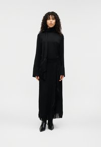 Μακρύ μαύρο φόρεμα με ζιβάγκο, κρόσσια στο τελείωμα και σχέδιο τύπου μπλούζας. Συνδυάζεται με ένα ταίριαστο κασκόλ, διαθέτει μακριά μανίκια και λεία ύφανση.