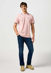 Jasnoróżowa koszulka polo z czarnym wykończeniem kołnierzyka, w zestawie z niebieskimi dżinsami i beżowymi mokasynami. Prosty design z małym emblematem na piersi.