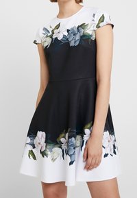 Robe sans manches noire avec un imprimé floral intégrant des accents blancs, bleus et verts au niveau de l'ourlet et du col, confectionnée en tissu lisse.