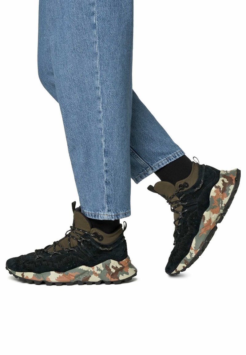 Chaussures de randonnée noires et vertes avec une tige texturée, des lacets solides et une semelle à motif camouflage multicolore. Portées avec un jean bleu retroussé.