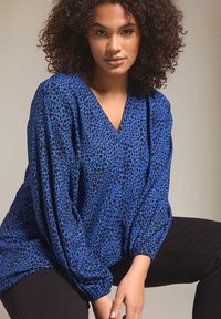 Blusa blu a stampa leopardata con scollatura a v, maniche lunghe a palloncino e polsini elasticizzati. Abbinata a pantaloni neri. Tessuto dalla consistenza liscia.