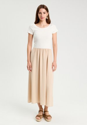 Valkoinen lyhythihainen paita, jossa on avara kaula-aukko, ja beige midi-hame. Pehmeä rakenne, valuva kangas ja tasapohjaiset sandaalit täydentävät asun.